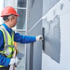 Budowlaniec w kasku i kamizelce nakłada tynk do elewacji na ścianę budynku za pomocą kielni.
