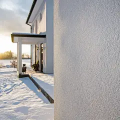 Śnieg otula dom z podkładem pod tynk silikonowy. Słońce przebija się przez chmury, oświetlając zimowy krajobraz.
