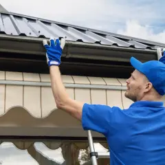 Mężczyzna w niebieskiej czapce i rękawiczkach montuje uszczelniacz do rynien plastikowych, dbając o szczelność dachu.